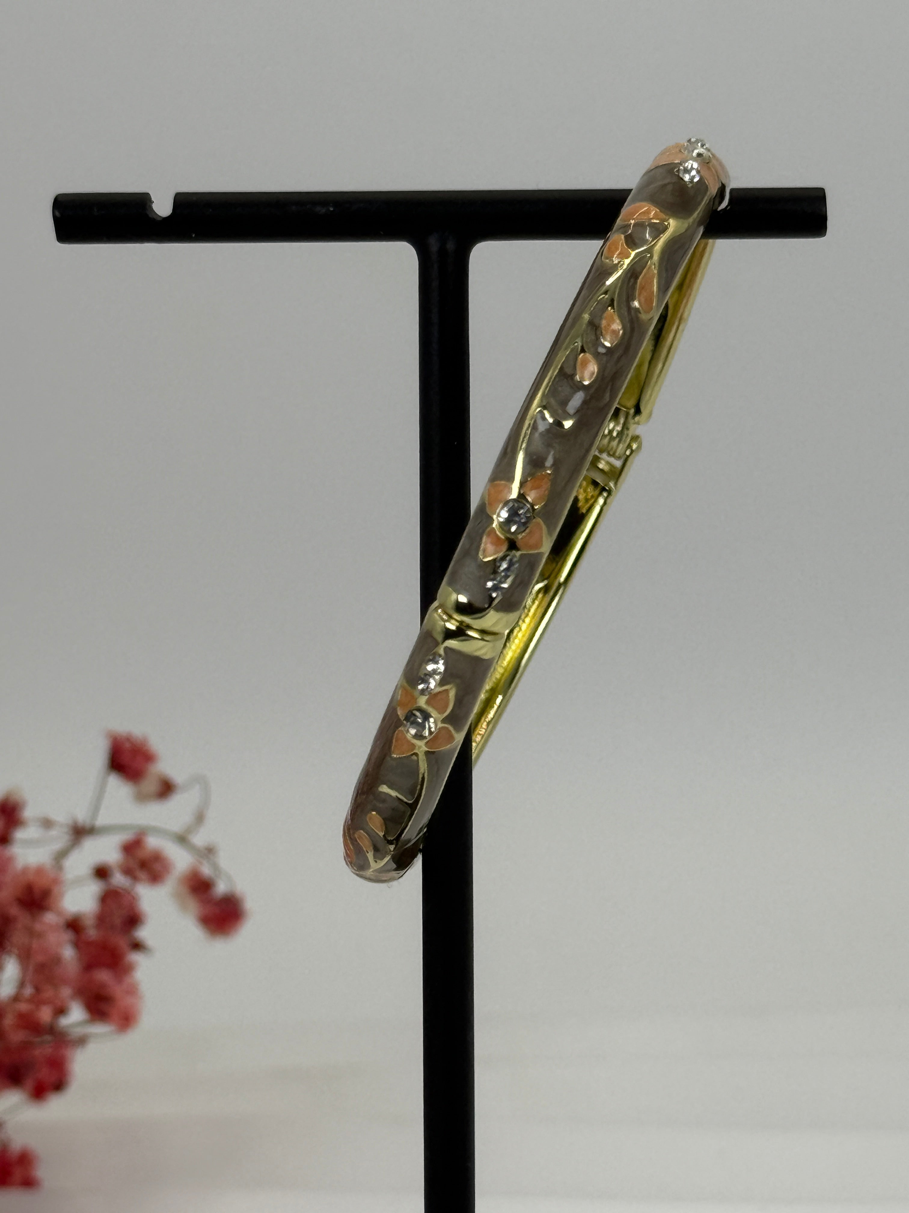 Opengaande messing gouden armband met grijs/taupe geëmailleerd bloemmotief – binnendiameter 6 cm.